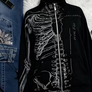 A black zip-up jacket with a white skeleton print on the front, next to blue jeans with white embroidery.