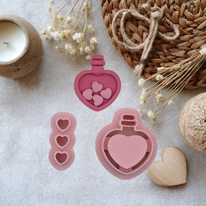 May include: A collection of pink heart-shaped cookie cutters and a wooden heart on a white surface. The cutters include a bottle shape with hearts inside, a bottle shape with a heart, and a vertical row of three hearts.