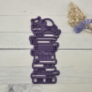 May include: A purple cat cookie cutter resting on a stack of books. The cat is curled up on top of the books. The books have various shapes and designs, including numbers and arrows.