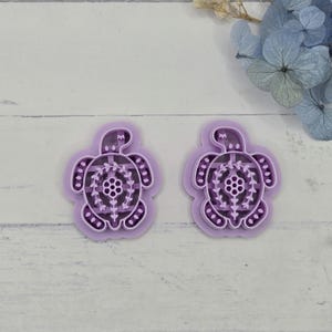 May include: Two lavender-colored turtle-shaped cookie cutters. Each cutter has a detailed design on the shell, with a central floral pattern and dotted edges. The cutters are on a light-colored wooden surface.