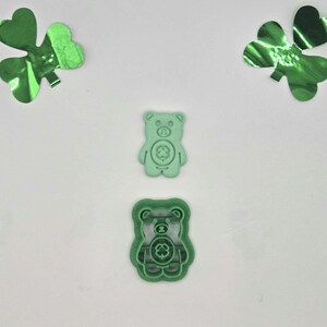 May include: Two green bear-shaped cookie cutters with a shamrock design, one with a cutting edge. Two green shamrock decorations are on a white background. The bear cutters are for St. Patrick's Day baking.