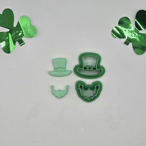 May include: A collection of four green St. Patrick's Day-themed cookie cutters: a top hat, a leprechaun hat, a beard, and a face. Two green shamrock decorations are in the background. The cutters are made of plastic and are on a white background.