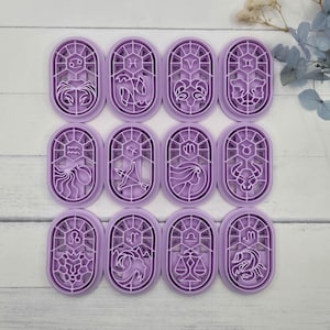 May include: A set of twelve oval-shaped lavender plastic cookie cutters, each featuring a unique zodiac sign symbol and corresponding astrological illustration within a stained-glass style frame, arranged in a grid on a white wooden surface.