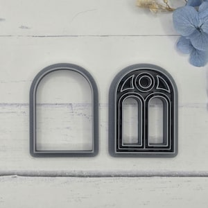 May include: Two gray plastic cookie cutters in the shape of arched windows. One is a simple arch, and the other has a detailed window design with two panes and an arched top. The cutters are on a white surface.