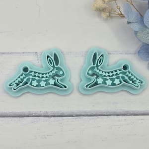 May include: Two light blue plastic bunny-shaped cookie cutters. Each cutter features a running rabbit design with star and floral cutouts. The cutters are on a light wood-grain surface, with dried flowers in the background.