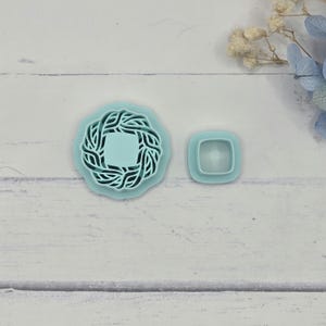 May include: Two light blue plastic cookie cutters. One is a square with rounded corners, and the other is a wreath shape with a square center. The wreath cutter has a detailed, woven design.