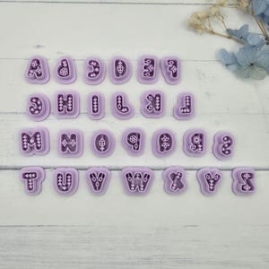May include: A set of purple alphabet cookie cutters, each with a decorative floral design. The letters are arranged in rows, showcasing the entire English alphabet. The cutters are made of plastic.