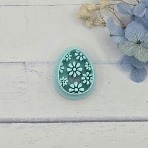 May include: A light blue, egg-shaped cookie cutter with a floral design. The cutter features a series of daisy-like flowers cut out of the center. The cutter is made of plastic and is designed for baking.