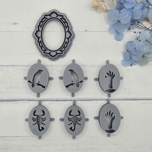 May include: A collection of gray, oval-shaped pendants and a decorative frame. The pendants feature cut-out designs of a bird on a branch, a hand, and a scorpion. The frame is oval with a decorative border.