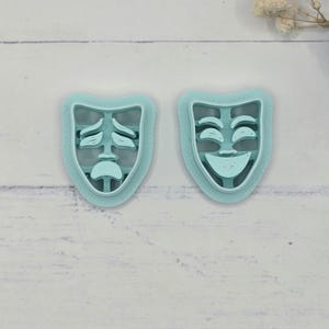 May include: Two light blue cookie cutters shaped like theater masks, one sad and one happy. The cutters are made of plastic and have a detailed design. These are ideal for creating cookies or other baked goods with a theatrical theme.