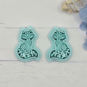 May include: Two teal fox-shaped craft embellishments with intricate white floral and leaf designs. The foxes have detailed faces and tails, perfect for crafting projects. The background is a light, neutral color.