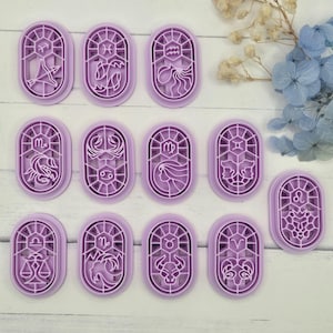 May include: A collection of twelve purple plastic cookie cutters, each featuring a different zodiac sign symbol within an oval frame. The designs include various astrological symbols, perfect for themed baking projects. The cutters are arranged on a light-colored surface.