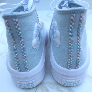 May include: Pair of light blue Converse All Star high-top sneakers. The sneakers have white soles and laces. The sides are embellished with rows of small, colorful beads. The Converse logo is visible on the side.