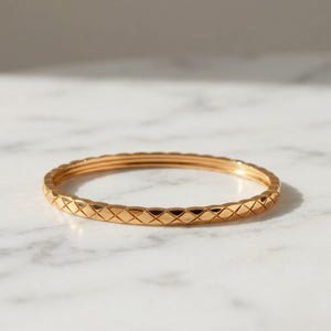 Puede incluir: Una pulsera de oro fina que presenta un patrón acolchado en forma de diamante que se repite en toda su superficie exterior. El metal tiene un acabado pulido y reflectante que capta la luz, creando una textura consistente en el aro.