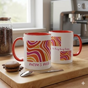 Könnte beinhalten: Zwei weiße Keramikbecher mit roten Griffen und Innenräumen. Auf einem Becher steht "mom's MUG" und auf dem anderen "I'm busy being happy". Beide Becher haben ein farbenfrohes abstraktes Design in Orange, Pink und Gelb.