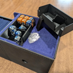 May include: A black gaming card storage box with a purple interior lining. The box features a removable black plastic dice tray, a compartment filled with orange and blue dice, and a silver emblem of a dragon head on a shield on the base.