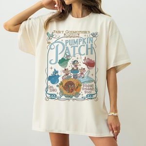 May include: Cream-colored t-shirt featuring a whimsical design from "Cinderella." The graphic includes the text "Fairy Godmother's Enchanted Pumpkin Patch" with illustrations of characters and a pumpkin carriage. The shirt also has the text "Est. 1980."