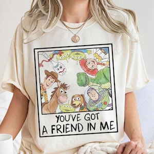 May include: Cream-colored t-shirt featuring a framed watercolor illustration of characters from the animated film "Toy Story." The image includes Woody, Buzz Lightyear, and other characters. The text "YOU'VE GOT A FRIEND IN ME" is printed below the illustration.