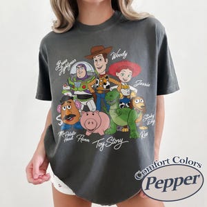 Puede incluir: Camiseta gris oscuro con un gr&aacute;fico de personajes de Toy Story, incluyendo a Woody, Buzz Lightyear y Jessie. La camiseta tiene las palabras "Toy Story" en una fuente de escritura. La camiseta tambi&eacute;n tiene las palabras "Comfort Colors Pepper" en un c&iacute;rculo.