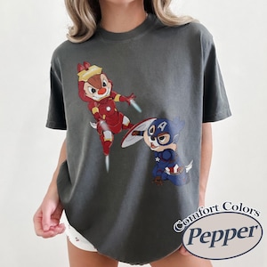 May include: Dark gray t-shirt featuring cartoon characters dressed as superheroes. One character is in a red Iron Man suit, and the other is in a Captain America suit. The shirt has the words "Comfort Colors Pepper" in a circular design.