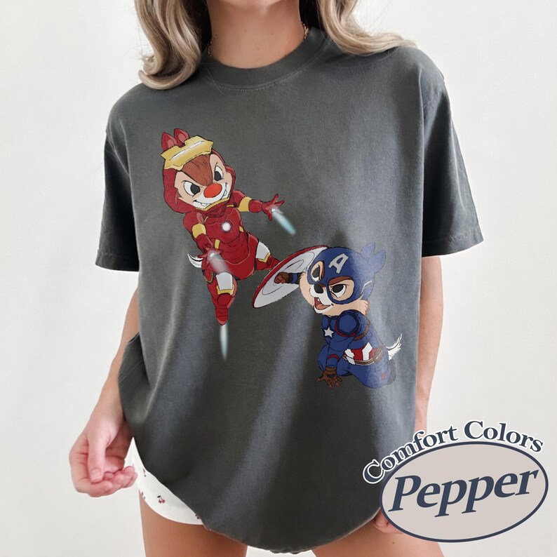 May include: Dark gray t-shirt featuring cartoon characters dressed as superheroes. One character is in a red Iron Man suit, and the other is in a Captain America suit. The shirt has the words "Comfort Colors Pepper" in a circular design.