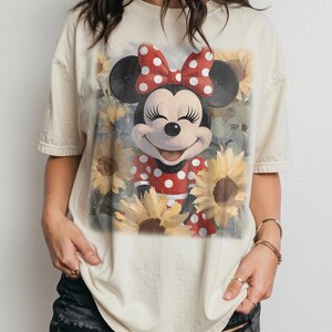 Puede incluir: Una camiseta de manga corta de color crema con un estampado gr&aacute;fico de Minnie Mouse con un vestido de lunares rojos y blancos y un lazo a juego, rodeada por un campo de girasoles amarillos con un acabado art&iacute;stico suave.