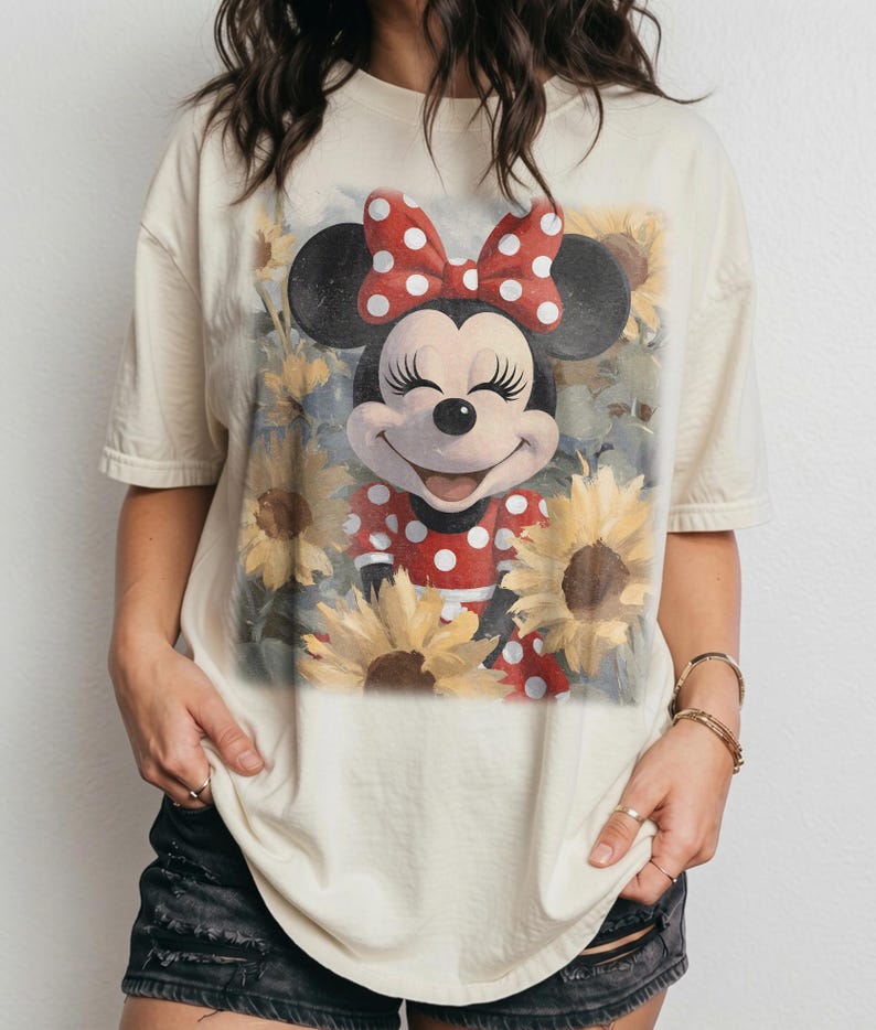 Puede incluir: Una camiseta de manga corta de color crema con un estampado gr&aacute;fico de Minnie Mouse con un vestido de lunares rojos y blancos y un lazo a juego, rodeada por un campo de girasoles amarillos con un acabado art&iacute;stico suave.