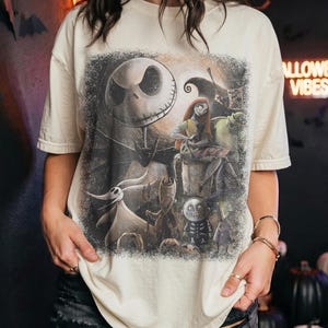 May include: A cream-colored short-sleeve t-shirt featuring a large, detailed graphic print of characters from The Nightmare Before Christmas, including Jack Skellington, Sally, Zero, and other figures, set against a dark, textured background.