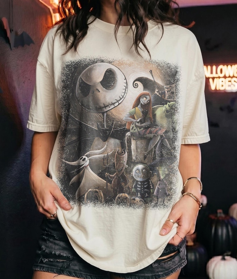 May include: A cream-colored short-sleeve t-shirt featuring a large, detailed graphic print of characters from The Nightmare Before Christmas, including Jack Skellington, Sally, Zero, and other figures, set against a dark, textured background.