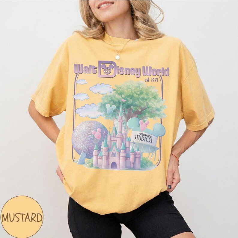 Puede incluir: Camiseta amarilla mostaza con un gr&aacute;fico de Walt Disney World, establecida en 1971. El dise&ntilde;o incluye un castillo, Spaceship Earth, nubes y el letrero de Hollywood Studios. La camiseta es de corte holgado.