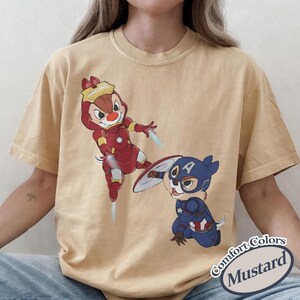 May include: Mustard-colored t-shirt featuring cartoon characters dressed as Iron Man and Captain America. The Iron Man character is in a red suit with yellow accents, while Captain America is in a blue suit with a shield. The shirt has the text "Comfort Colors Mustard".