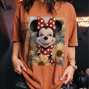 Puede incluir: Una camiseta de color naranja quemado con un estampado gr&aacute;fico de Minnie Mouse con un vestido de lunares rojos y blancos y un lazo a juego, rodeada de girasoles amarillos. El personaje tiene una expresi&oacute;n sonriente con ojos cerrados.