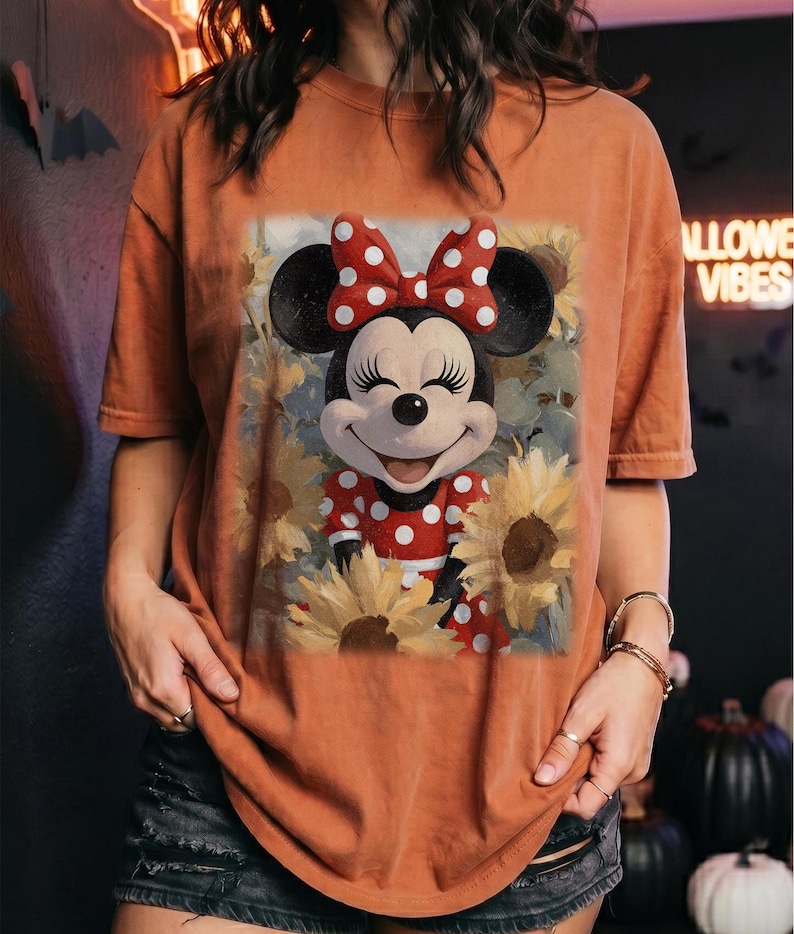 Puede incluir: Una camiseta de color naranja quemado con un estampado gr&aacute;fico de Minnie Mouse con un vestido de lunares rojos y blancos y un lazo a juego, rodeada de girasoles amarillos. El personaje tiene una expresi&oacute;n sonriente con ojos cerrados.