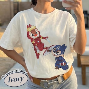 May include: Ivory-colored t-shirt featuring cartoon characters dressed as superheroes. One character is in a red Iron Man suit, and the other is in a Captain America costume. The shirt has the words "Comfort Colors" and "Ivory" printed on it.