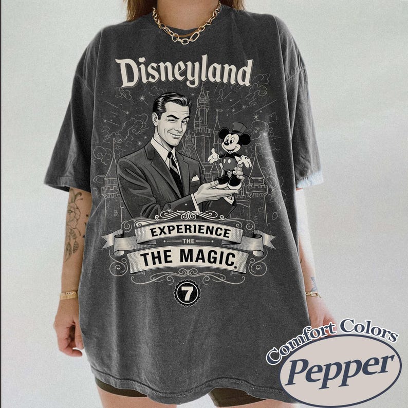 Pu&ograve; includere: T-shirt grigio scuro con grafica vintage di Disneyland. Il design include un uomo che tiene in mano una figura di Topolino, la scritta "Disneyland" e "Experience the Magic". La maglietta ha un'etichetta Comfort Colors Pepper.