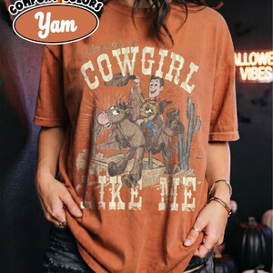 May include: A yam-colored short-sleeve t-shirt featuring a graphic print of a cowboy riding a horse in a desert landscape with cacti. The text reads "You're a COWGIRL LIKE ME" in a distressed font. The shirt has a crew neckline and a relaxed fit.