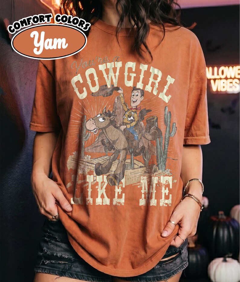May include: A yam-colored short-sleeve t-shirt featuring a graphic print of a cowboy riding a horse in a desert landscape with cacti. The text reads "You're a COWGIRL LIKE ME" in a distressed font. The shirt has a crew neckline and a relaxed fit.
