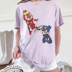 May include: A light purple t-shirt featuring cartoon characters dressed as superheroes. One character is in a red Iron Man suit, and the other is in a Captain America suit. The shirt has the words "Comfort Colors Orchid" on a label.