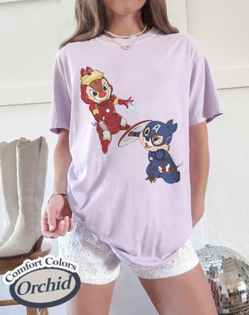 May include: A light purple t-shirt featuring cartoon characters dressed as superheroes. One character is in a red Iron Man suit, and the other is in a Captain America suit. The shirt has the words "Comfort Colors Orchid" on a label.