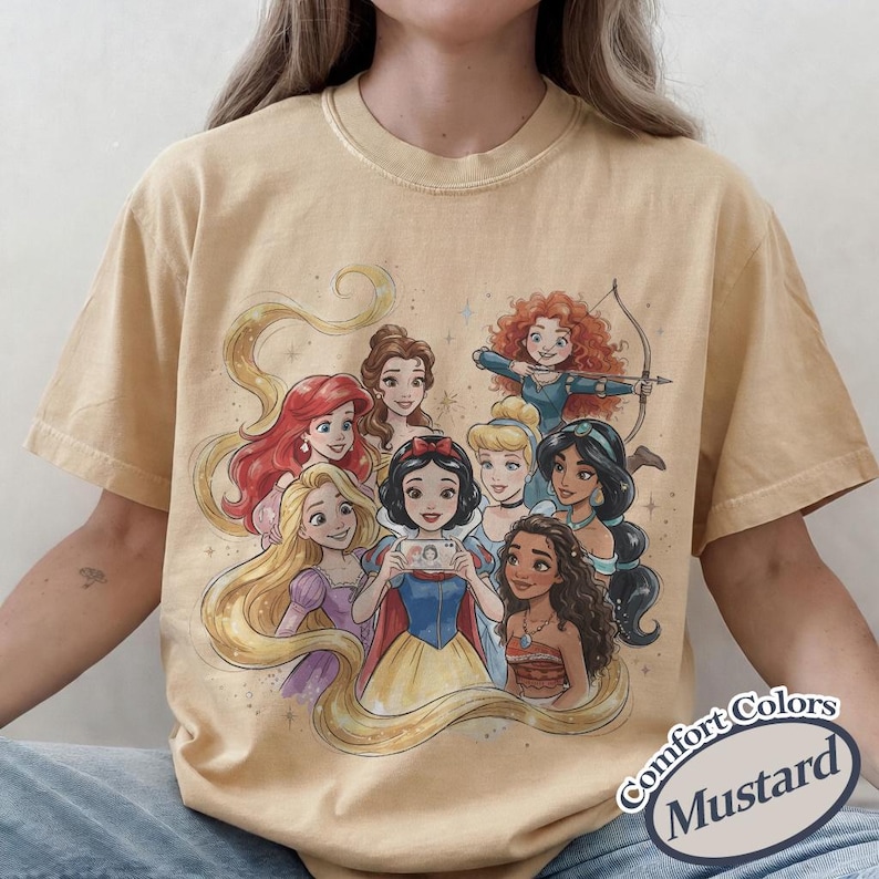 Op de afbeelding: Mosterdkleurig T-shirt met een afbeelding van acht geanimeerde prinsessen. De prinsessen worden afgebeeld met verschillende kapsels en outfits. Het shirt heeft een relaxte pasvorm en de woorden "Comfort Colors" en "Mustard" staan rechtsonder.