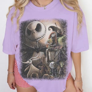 May include: A light purple short-sleeve t-shirt featuring a large, detailed graphic print of characters from The Nightmare Before Christmas, including Jack Skellington, Sally, Zero, and the trick-or-treaters, set against a dark, textured background.
