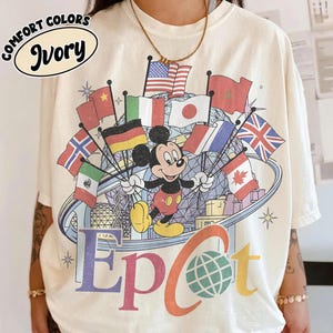 May include: An ivory t-shirt featuring a graphic of Mickey Mouse standing in front of the Epcot geodesic sphere. The design includes various international flags, the text "EpCot" with a globe icon, and a "COMFORT COLORS Ivory" label.