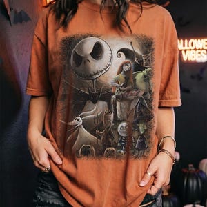 May include: A burnt orange t-shirt featuring a detailed graphic print of characters from The Nightmare Before Christmas, including Jack Skellington, Sally, Zero, and the trick-or-treaters, set against a dark, textured, and moody background.