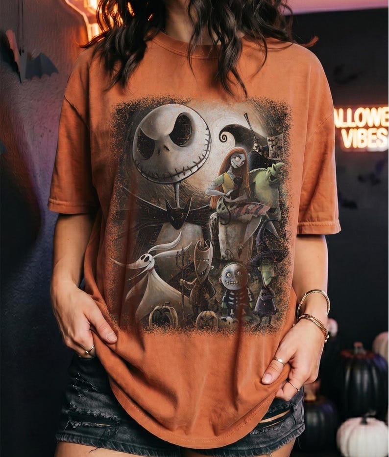 May include: A burnt orange t-shirt featuring a detailed graphic print of characters from The Nightmare Before Christmas, including Jack Skellington, Sally, Zero, and the trick-or-treaters, set against a dark, textured, and moody background.