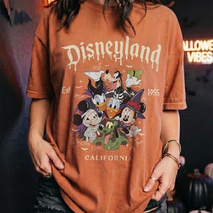 May include: A burnt orange short-sleeve t-shirt featuring a graphic of Disney characters in Halloween costumes. The text reads Disneyland Est 1955 California. The characters include Mickey Mouse as a mummy, Minnie Mouse as a witch, and Goofy.