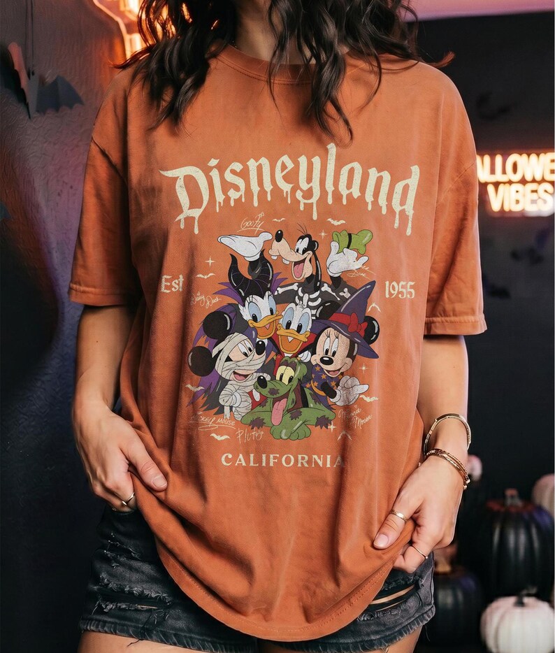 May include: A burnt orange short-sleeve t-shirt featuring a graphic of Disney characters in Halloween costumes. The text reads Disneyland Est 1955 California. The characters include Mickey Mouse as a mummy, Minnie Mouse as a witch, and Goofy.