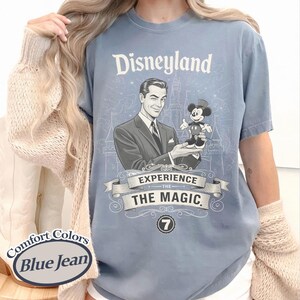 Pu&ograve; includere: T-shirt blu jeans Comfort Colors con grafica Disneyland. La grafica presenta un uomo in giacca e cravatta che tiene Topolino, con la scritta "Disneyland" e "Experience the Magic". &Egrave; presente anche il numero 7.