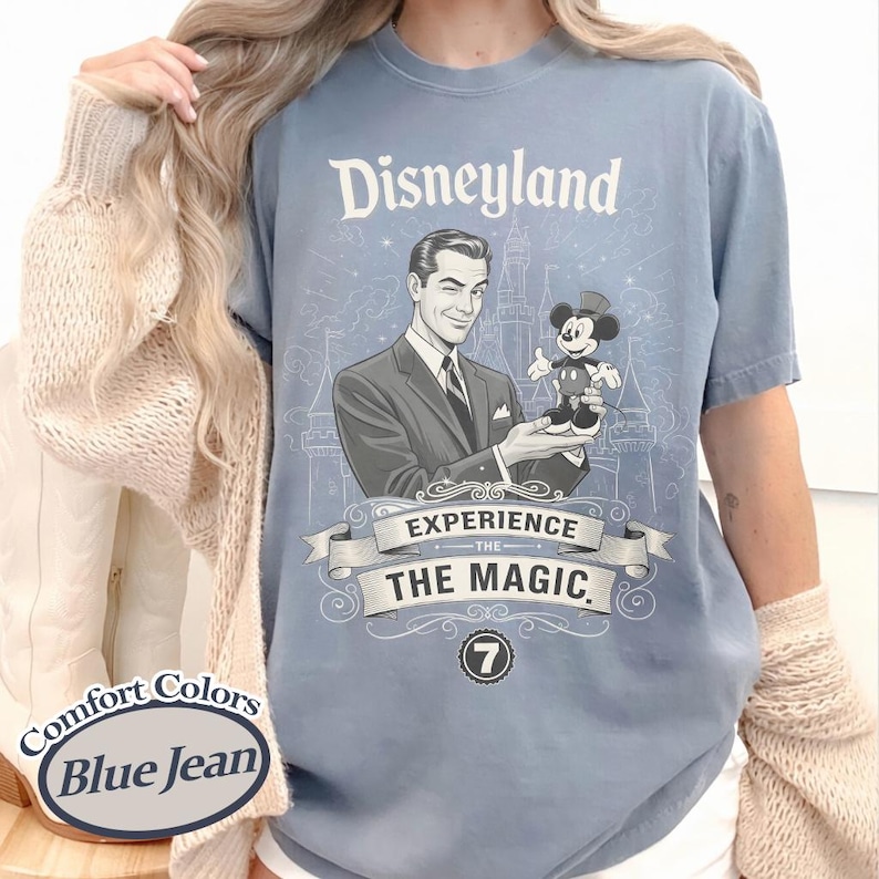Pu&ograve; includere: T-shirt blu jeans Comfort Colors con grafica Disneyland. La grafica presenta un uomo in giacca e cravatta che tiene Topolino, con la scritta "Disneyland" e "Experience the Magic". &Egrave; presente anche il numero 7.