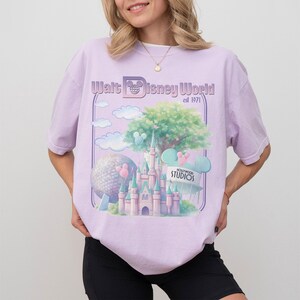 Puede incluir: Camiseta lila claro con un gr&aacute;fico del castillo de Cenicienta, un &aacute;rbol y una gran esfera. Tambi&eacute;n es visible el texto "Walt Disney World est 1971" y "Hollywood Studios". La camiseta es de corte holgado.