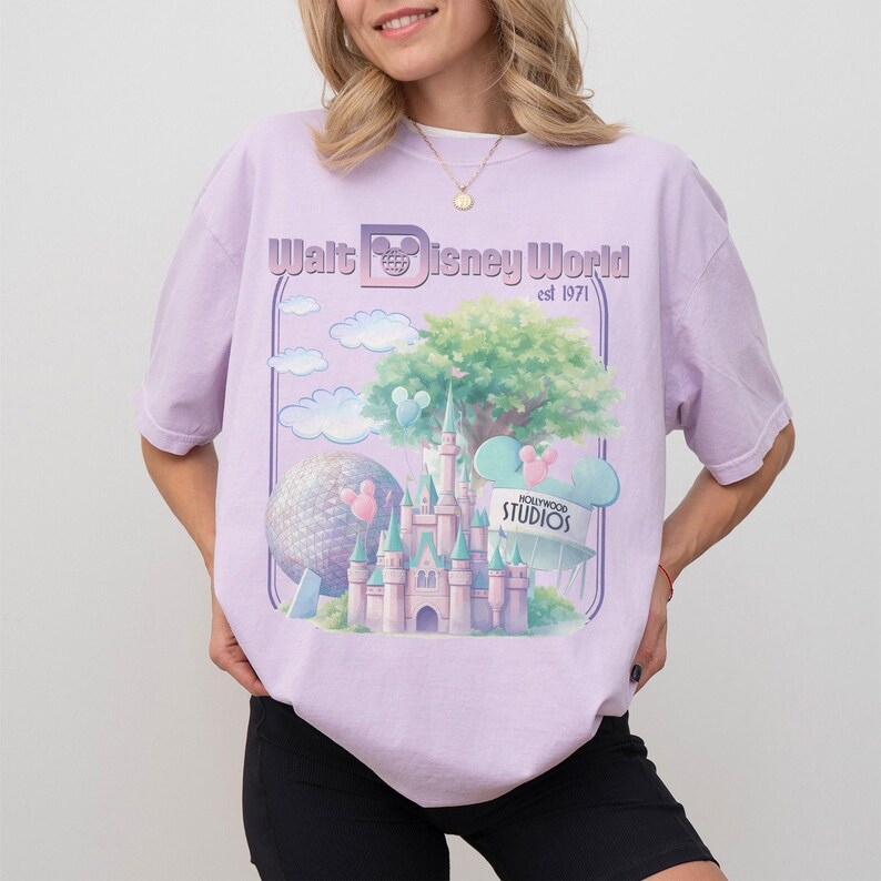 Puede incluir: Camiseta lila claro con un gr&aacute;fico del castillo de Cenicienta, un &aacute;rbol y una gran esfera. Tambi&eacute;n es visible el texto "Walt Disney World est 1971" y "Hollywood Studios". La camiseta es de corte holgado.